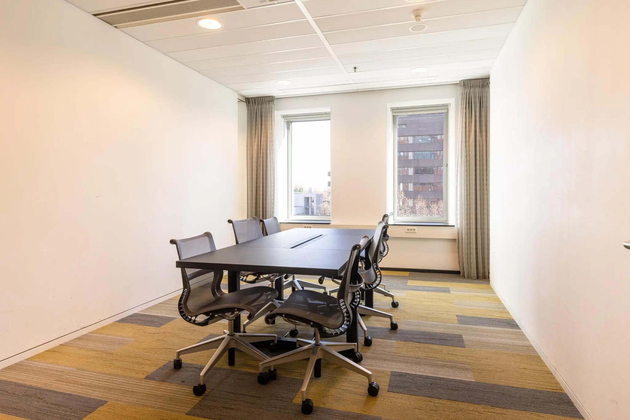 Kleine vergaderruimte aan de Prinses Beatrixlaan met zes stoelen rond een tafel en uitzicht op kantoorgebouwen.